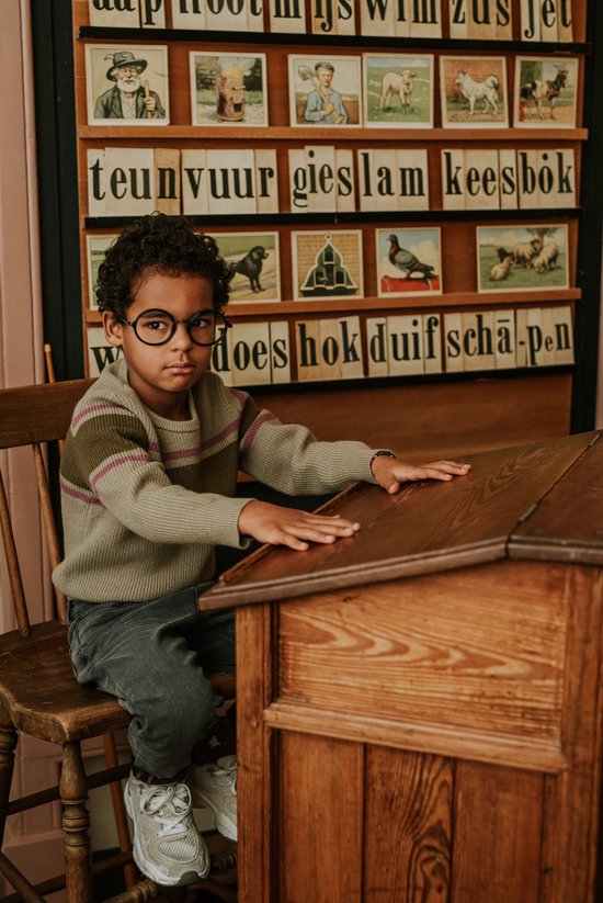 Jongen met krullend haar en bril, zittend aan een houten schoolbureau, draagt een gestreepte trui en grijze jeans.
