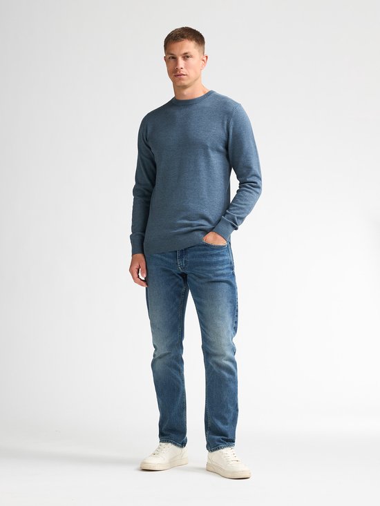 Een man staat met zijn handen in zijn zakken, gekleed in een blauwe, fijngebreide trui en spijkerbroek.