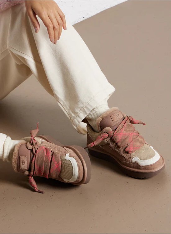 Vrouw met bruine UGG sneakers met roze veters, gedragen met een beige broek.