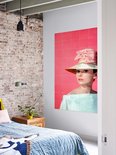 Audrey Hepburn portant un grand chapeau, un nœud rose et un haut bleu pâle, sur fond rose, dans une chambre avec mur de briques.