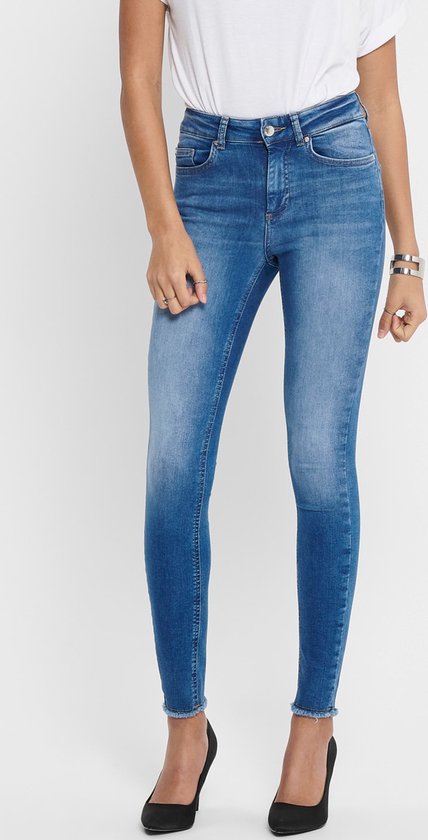 Model draagt een skinny jeans met blauwe denim wassing, rafelige zoom en zwarte pumps. Gedetailleerde sieraden zichtbaar.