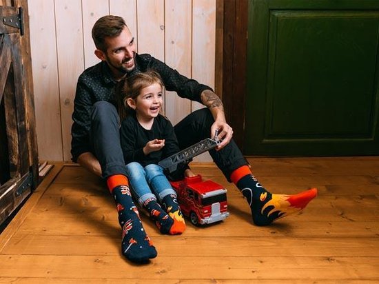 Vrolijk kind en volwassene zitten op een houten vloer en dragen blauwe sokken met brandweerwagens en vlammen.