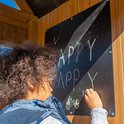 Kind met krullend haar schrijft 'HAPPY' op een zwart schoolbord op een houten speeltoestel.