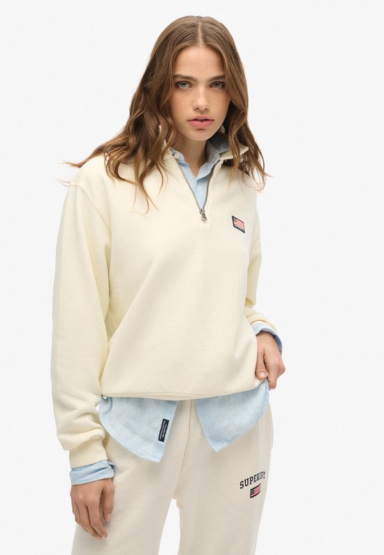 Model draagt een off-white trui met halve rits, lichtblauwe gestreepte blouse eronder en een patch op de borst.
