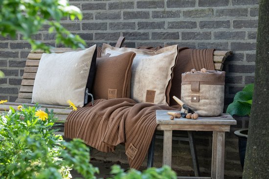Bruine gebreide plaid met geribd patroon op een houten bankje met bruine en beige kussens.