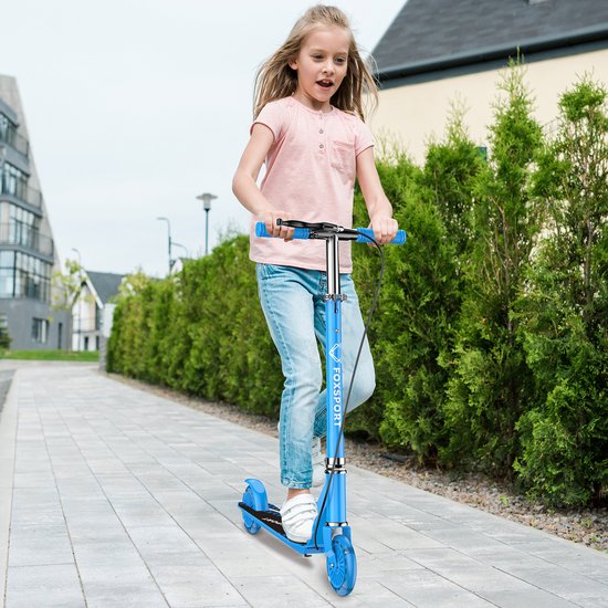 Meisje met blonde haren die op een blauwe step rijdt op een betegeld pad, met groen struikgewas op de achtergrond.
