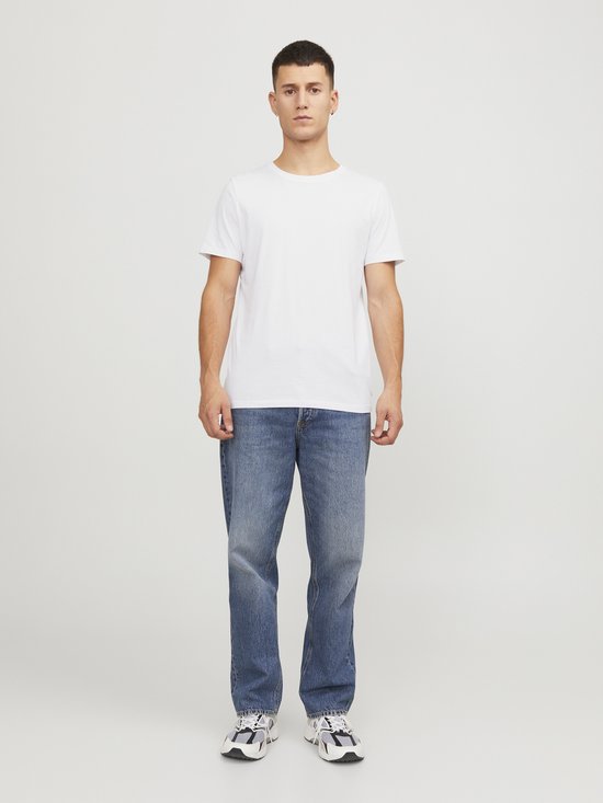 Man in een wit T-shirt en blauwe spijkerbroek, staand met de handen langs het lichaam.