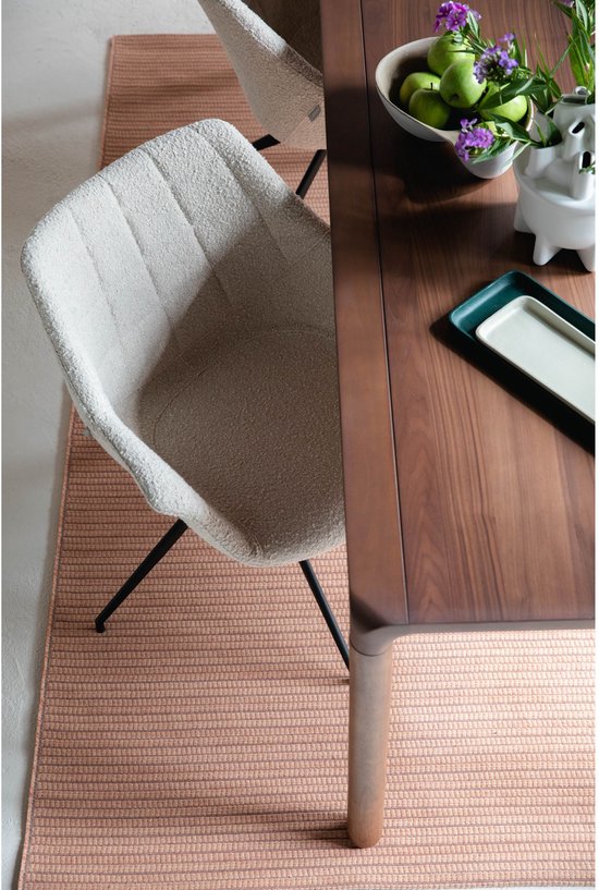 Gedeelte van een bruine walnoot eettafel met beige fauteuil met metalen poten, staand op een roze vloerkleed.