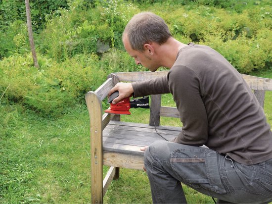 Man schuurt een houten bankje in de tuin met een rode Einhell multischuurmachine.