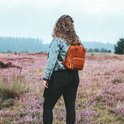Een vrouw met krullend haar, een spijkerjack en zwarte broek, draagt een bruine leren rugzak in een veld met paarse bloemen.