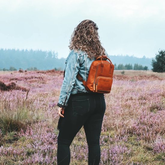 Een vrouw met krullend haar, een spijkerjack en zwarte broek, draagt een bruine leren rugzak in een veld met paarse bloemen.