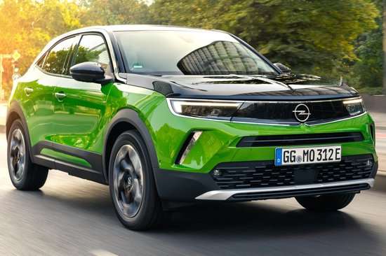 Groene Opel Mokka rijdt op een weg, met een stadsgebouw op de achtergrond.