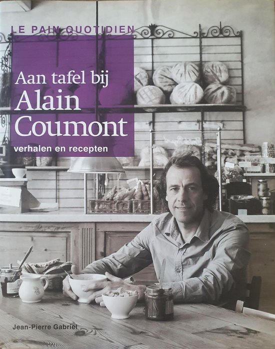 De man op de foto is Alain Coumont, zittend aan een tafel met een schaal met brood, potten jam en kommen ontbijtgranen. De achtergrond is een bakkerij met rekken vol brood.