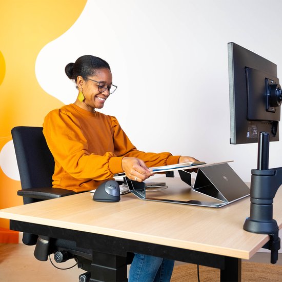 Vrouw met oranje trui en bril bekijkt documenten op een zwarte documenthouder aan een bureau.