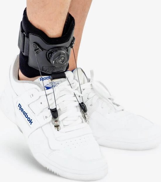 Detail van een zwarte klapvoetbrace met BOA-sluiting, bevestigd aan de veters van een witte Reebok sneaker.