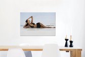 Canvas Schilderij Een vrouw in een zwarte bikini poseert op het strand - 60x40 cm - Wanddecoratie