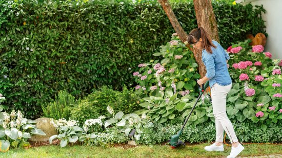 Vrouw in spijkerblouse trimt gras in een weelderige tuin met bloeiende planten met de Bosch grastrimmer.