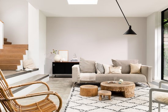 Lichte woonkamer met een stoffen bank, rotan stoel, houten salontafel en een open haard met trap.