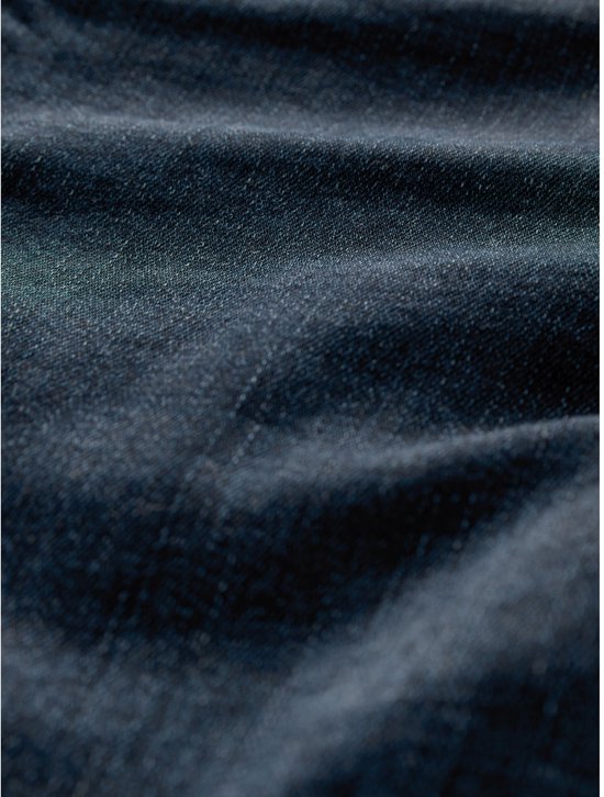 Detailopname van de textuur van een donkerblauwe spijkerstof. De stof is subtiel golvend en heeft een fijne weving.