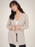 Vrouw met lange, golvende donkere haren in een lange, beige blazer met open voorkant en een witte top.