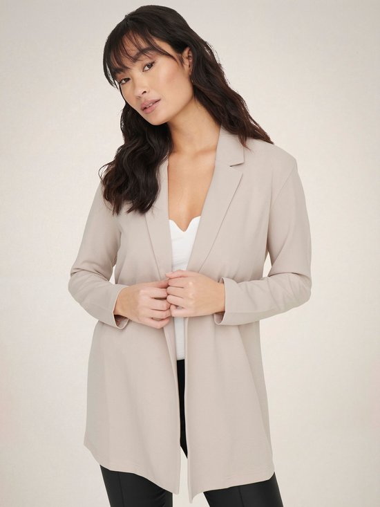 Vrouw met lange, golvende donkere haren in een lange, beige blazer met open voorkant en een witte top.