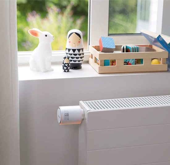 Netatmo slimme thermostaatknop, wit met oranje accent, op een witte radiator. Kinderkamer details op achtergrond.