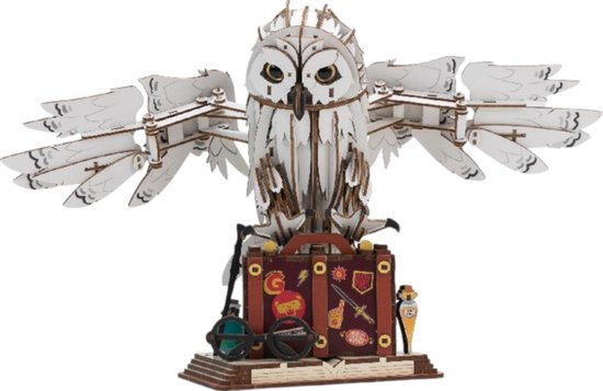 Houten model van Hedwig, de sneeuwuil, met uitgespreide vleugels, zittend op een koffer versierd met Harry Potter-symbolen. Staat op een open boek.