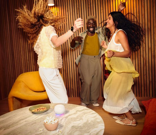 Drie mensen die dansen in een woonkamer, met de witte Google Home speaker op de salontafel.
