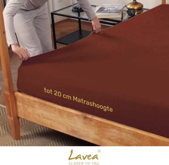 Vrouw trekt een kastanjebruin jersey hoeslaken met elastiek aan op een bed. Tekst: "tot 20 cm Matrashoogte".