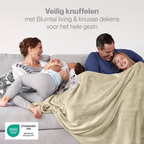Een gezin knuffelt samen onder een zandkleurige fleece deken op een grijze bank. OEKO-TEX gecertificeerd.