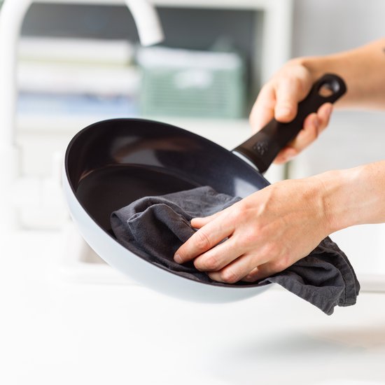 Handen die een lichtblauwe pan met zwarte binnenkant en grijze greep afdrogen.