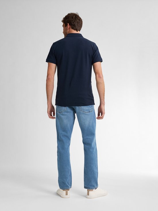 Man in donkerblauw poloshirt en lichtblauwe jeans, staand.