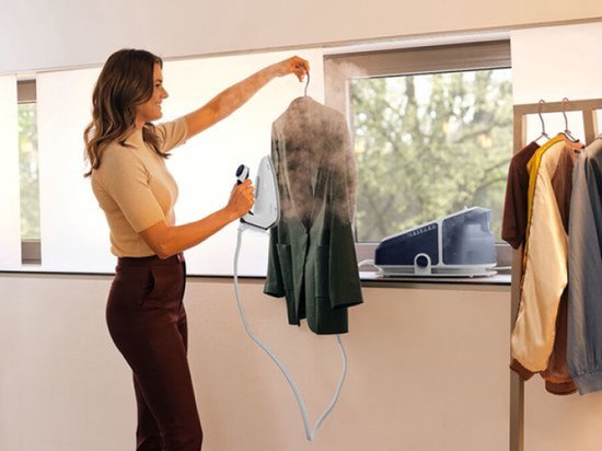 Vrouw gebruikt Braun CareStyle 7 Pro om jas te stomen, met kledingrek op de achtergrond.
