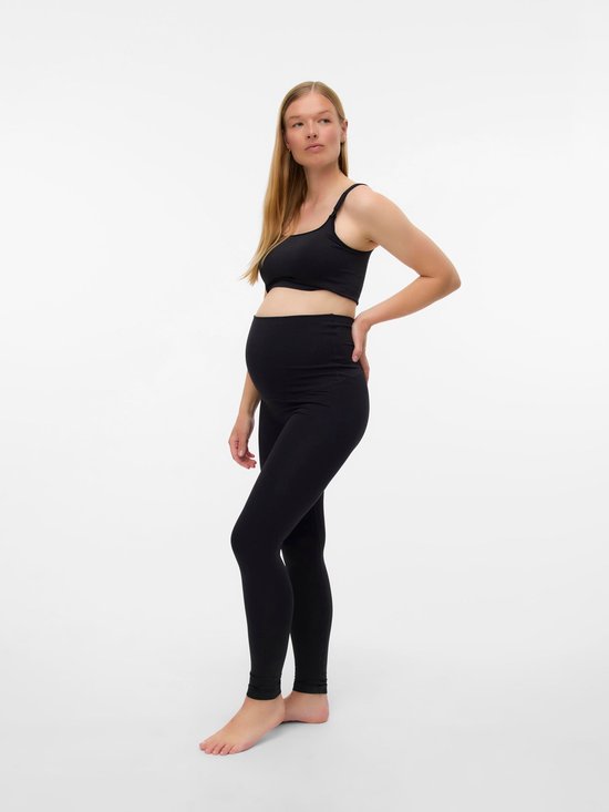 Zwanger model met blonde haren in een zwarte sportbeha en legging, hand op heup.