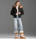 Model in een donkergroene, gewatteerde bomberjack met capuchon en bruine bontkraag. Draagt een witte coltrui en lichte jeans.