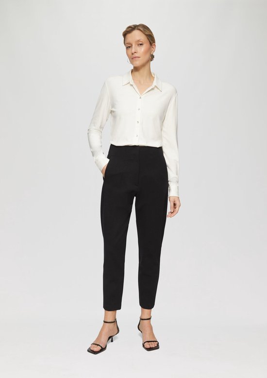 Model draagt een zwarte aansluitende tricot broek met taps toelopende pijpen en een witte blouse.