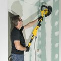 Man met veiligheidsbril schuurt een muur met gele en zwarte telescoopschuurmachine, aangesloten op een zwarte slang.