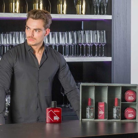 Man in donkergrijs overhemd staat naast een bar met Lock Stock & Barrel producten. Rode fles shampoo op de bar.