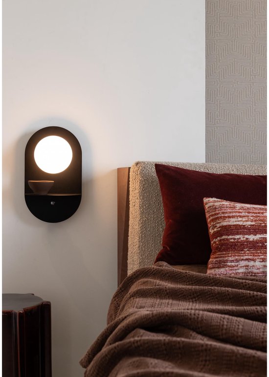 Zwarte wandlamp met ovale kap en klein rond schapje. Staat naast een bed met bruine deken en kussen.