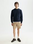 Model draagt een donkerblauwe sweater met ronde hals en beige short. Een witte T-shirt is zichtbaar onder de sweater.