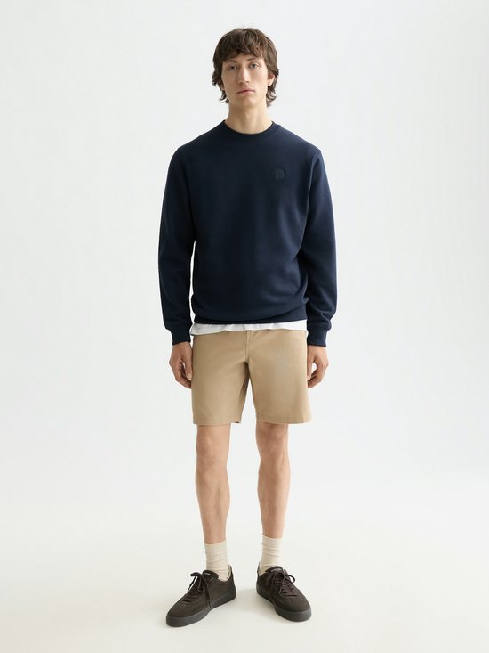 Model draagt een donkerblauwe sweater met ronde hals en beige short. Een witte T-shirt is zichtbaar onder de sweater.