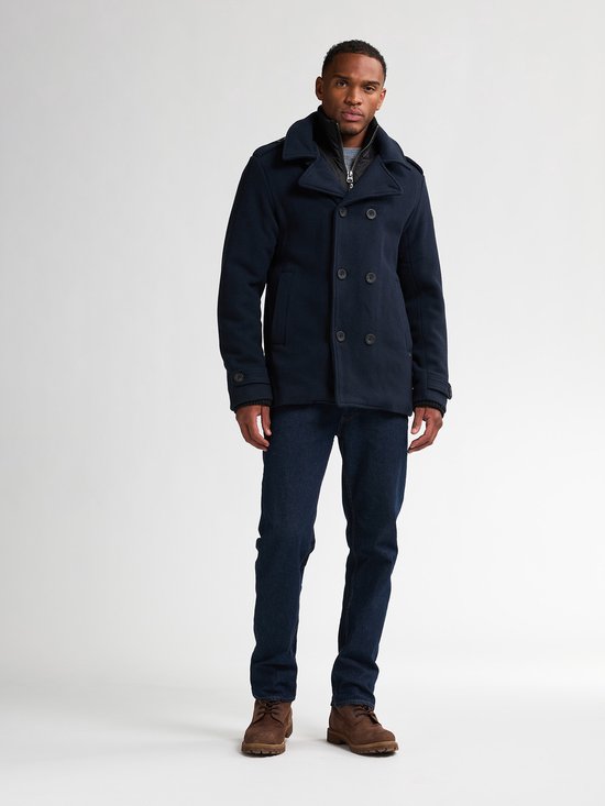 Man in donkerblauwe peacoat, jeans en bruine schoenen, staand.