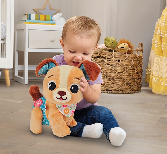 Een baby speelt met een zachte, bruine puppy met een lichtblauwe, bloemrijke broek en een bandana.