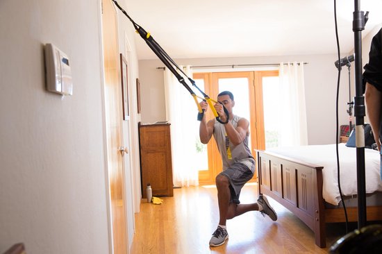 Man met een TRX-trainingsband en een sportshirt in een slaapkamer, doet een oefening op één knie.