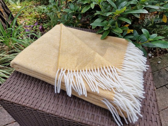 Okergele wollen deken met visgraatpatroon en witte franjes, op een bruine gevlochten bijzettafel in de tuin.