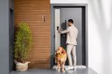 Man in beige pak ontgrendelt een donkergrijze deur met een zwarte deurbel; naast hem een golden retriever.