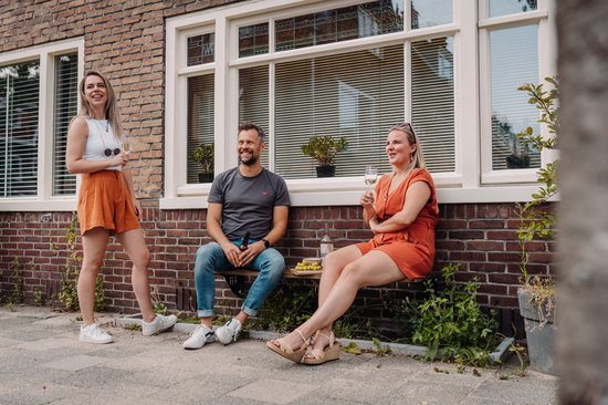 Mensen zitten en staan op houten gevelbank voor bakstenen muur, met drankjes.
