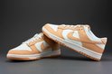 Een paar Nike Dunk Low damesschoenen, wit met oranje overlays, op een grijze achtergrond.