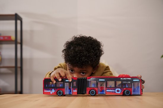 Jongen met krullend haar kijkt naar een rode gelede stadsbus met zwarte accenten.