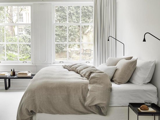 Lichtrijke slaapkamer met een opgemaakt bed met beige linnen beddengoed, grote ramen en zwarte lampen.